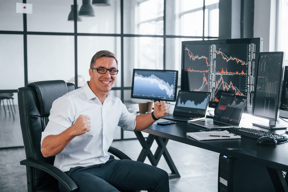 Man in a white shirt sits at a dark desk with multiple monitors showing stock charts, fists clenched in celebration.