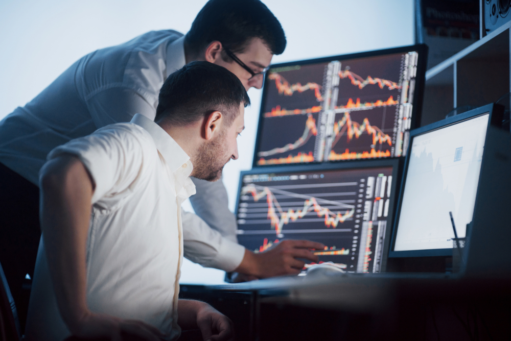 Two professionals monitor multiple stock charts on their screens, analyzing market data together in a dim office.